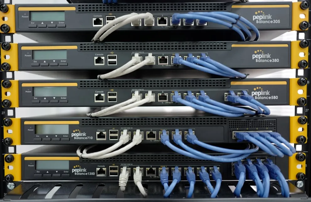 Licenza FIPS per Balance 310, cinque router Peplink montati in rack con cavi ethernet e schermi LCD in ambiente server.