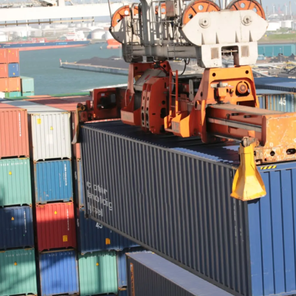 Orange port crane lifting a dark blue shipping container at a busy dockyard with stacked multicolored containers and ships