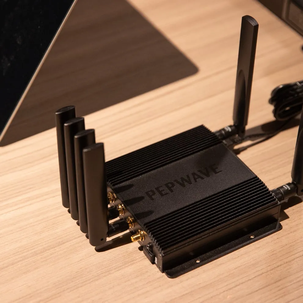 Black Pepwave TA Cloud Appliance router with finned metal casing, four left-side antennas, rear antenna, and mounting