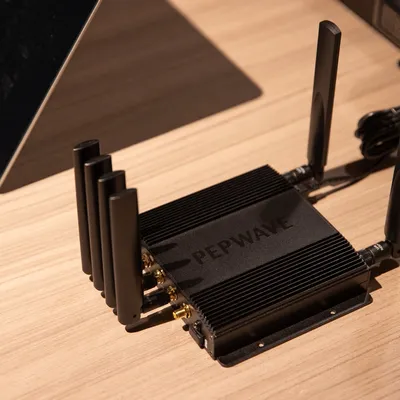 Dispositivo di rete Pepwave nero con dissipatore alettato e 5 antenne su superficie in legno chiaro accanto a un laptop
