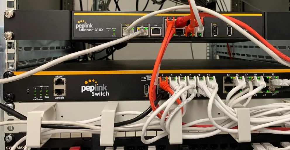 Router FIPS Balance 310 montato su rack sopra switch Peplink con cavi Ethernet collegati e LED verdi accesi.