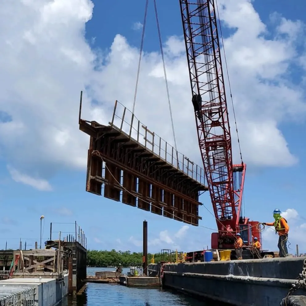 Red lattice crane lifting large steel bridge section over water during construction with workers on barge