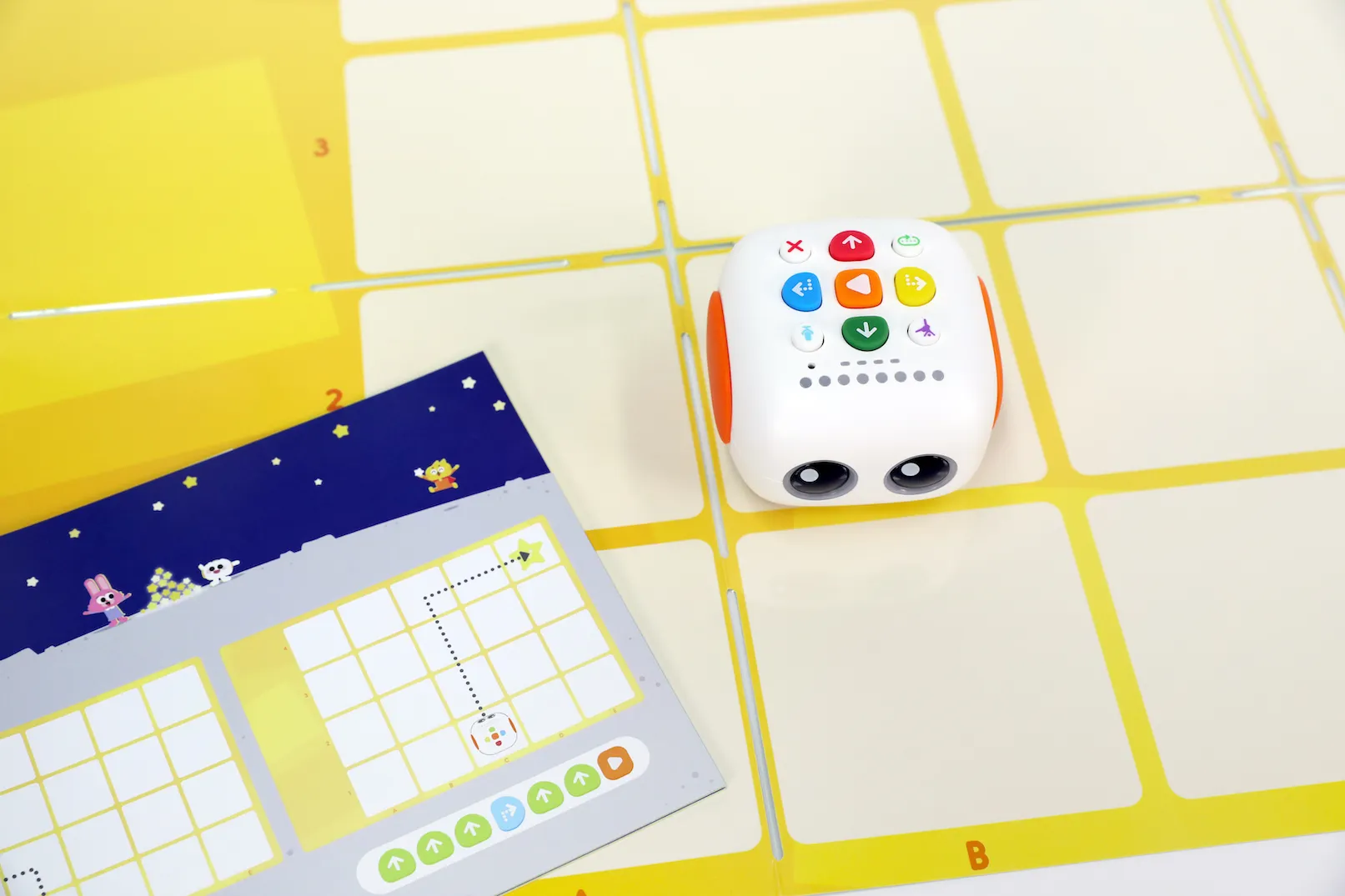 Matatastudio MatataLab MINT Roboter TaleBot Pro, white cube robot with orange wheels on yellow grid mat, featuring colorful Matatastudio MatataLab MINT Roboter TaleBot Pro, white cube robot with orange wheels on yellow grid mat, featuring colorful
