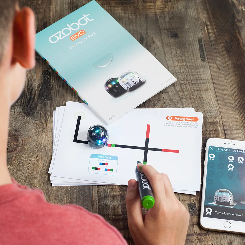 Ozobot MINT Coding Robot Evo Entry Kit, small dome-shaped robot with clear top and blue LEDs on printed track with child's Ozobot MINT Coding Robot Evo Entry Kit, small dome-shaped robot with clear top and blue LEDs on printed track with child's