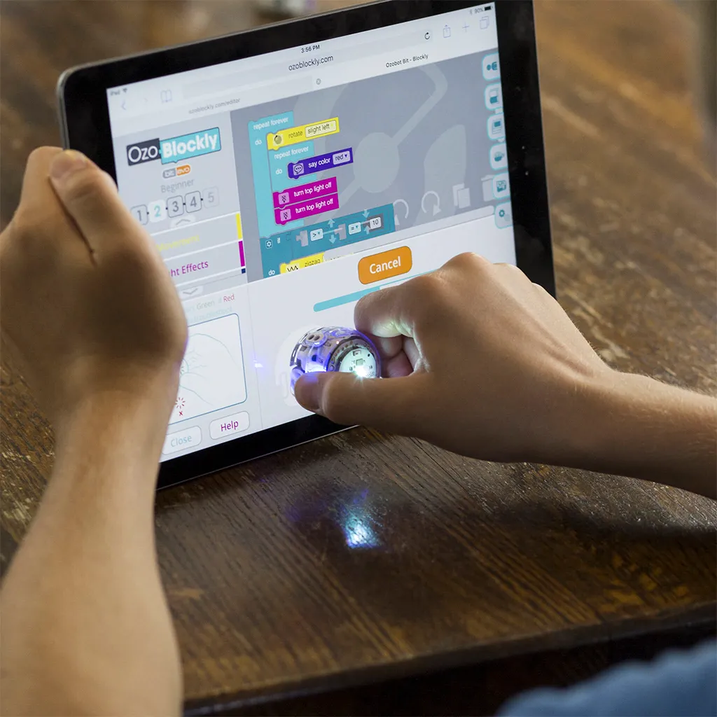 Ozobot MINT Coding Roboter Evo Entry Kit, person holding small round robot with blue lights against tablet screen showing Ozobot MINT Coding Roboter Evo Entry Kit, person holding small round robot with blue lights against tablet screen showing