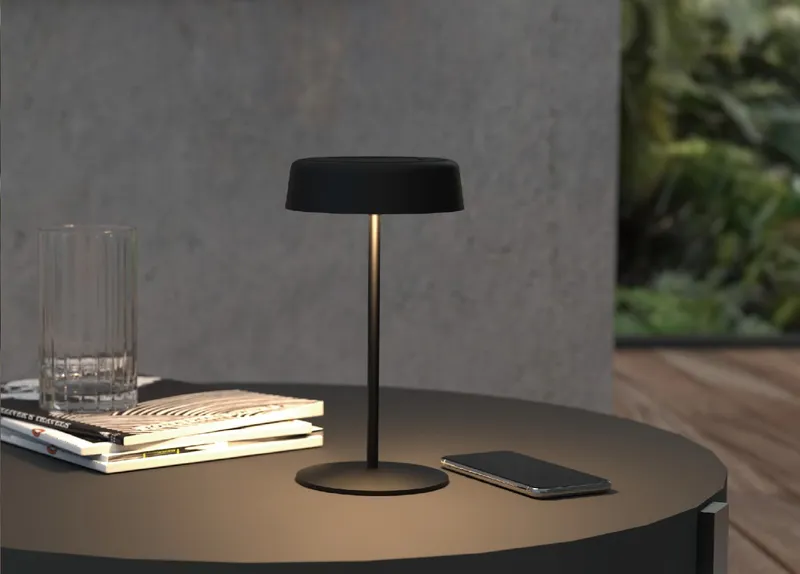 Lámpara de mesa negra Synergy 21 LED SOLAR con base circular, pantalla plana y luz cálida en mesa exterior junto a libros y